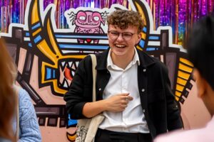 Matthew stands infront of a large character print out of an illustration that he has done. He is wearing a white shirt and black jacket, and are laughing to people out of shot