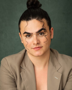 Joey is looking towards the camera in a headshot style photo. their hair is tied back in a bun, two strands of hair fall down the front of their face