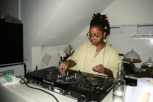 Iesha Palmer, a woman with afro-textured hair stands wearing headphones and DJing at some decks