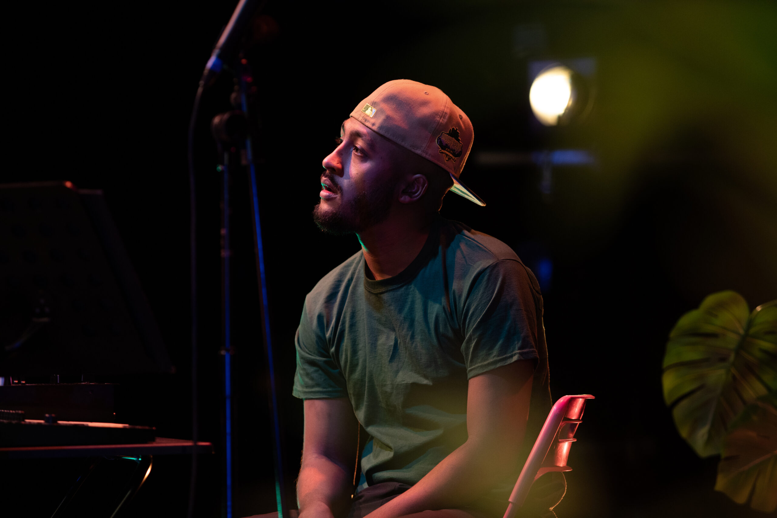Mal is wearing a peach coloured snapback cap backwards. He is also wearing a green t-shirt. He sits in a studio on a pink chair and appears to be listening to somebody talking who is outside of the frame of this image.