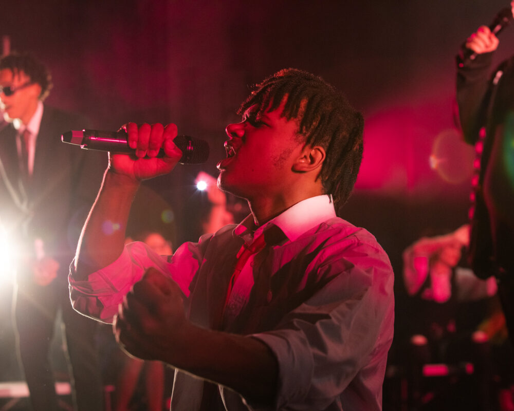 A male wearing a white shirt raps into a mic. The lighting is dim and red.