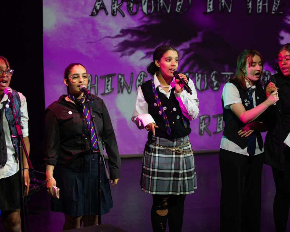 A group of five females wearing punk uniforms sing into mics