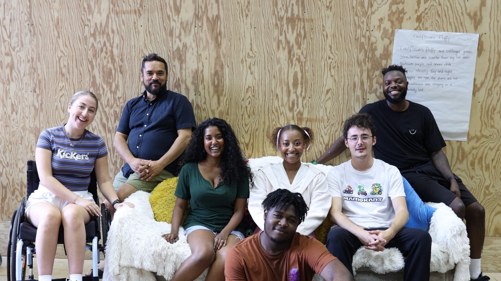 A group of people sit smiling facing the camera. they are on or around a sofa.