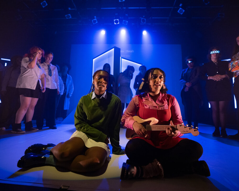 Two Black females sit on stage. One is playing a ukelele. The lighting on stage is blue.
