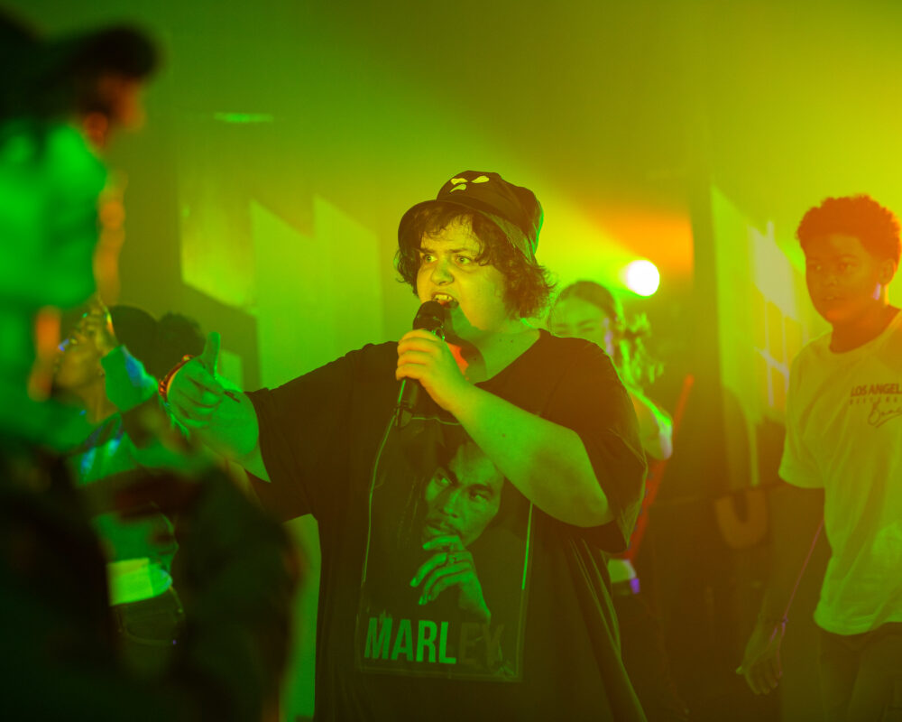 a young white male wearing a bucket hat raps into a mic. the lighting is atmospheric and green/yellow
