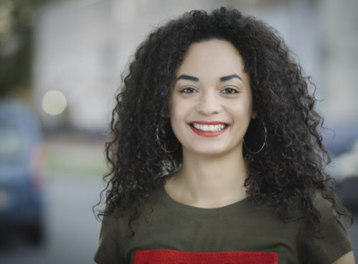 Anita is standing on a street wearing a khaki green tshirt with red details. She has long, curly black hair and a bright smile. She is wearing red lipstick and large silver hoop earrings. 
