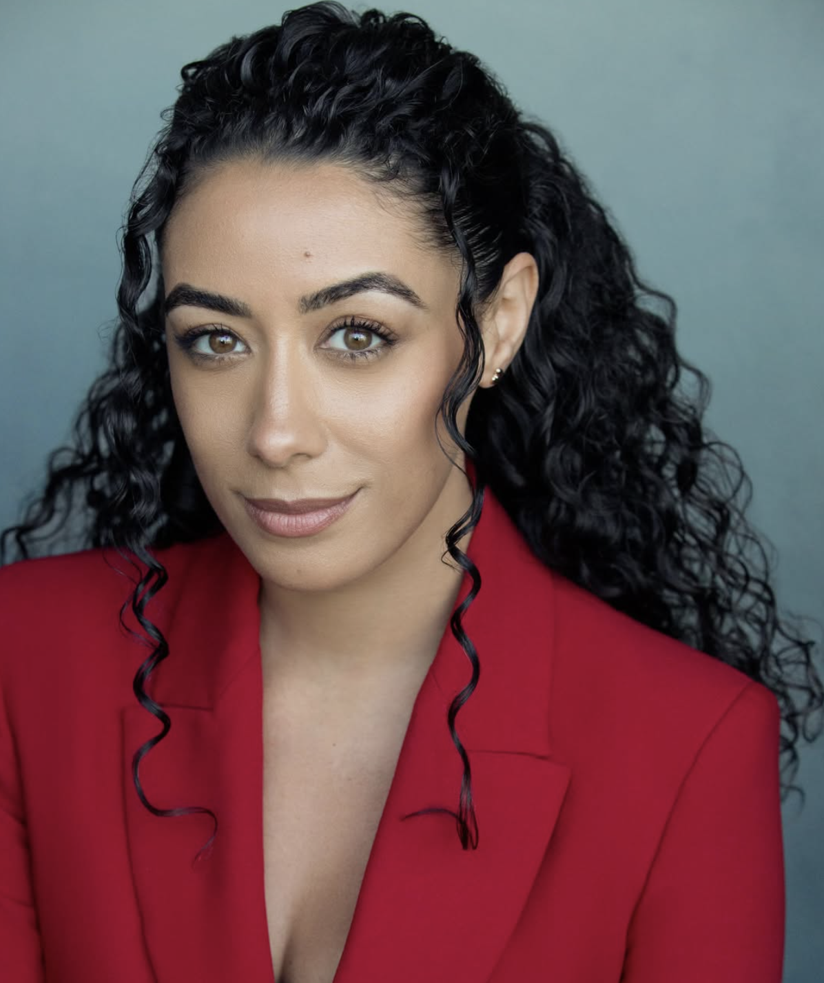 Paislie sits against a plain blue background. She has long curly black hair. She is wearing small gold stud earrings and a red blazer. 