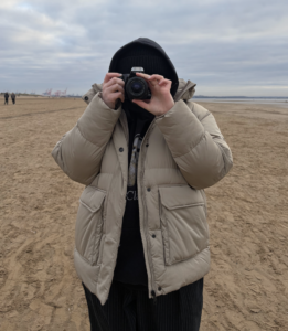 AJ stands holding a camera, they take a picture of the person who is taking a picture of them. They are wearing a beige puffer jacket and appear to be stood on a beach, with clouds in shot.
