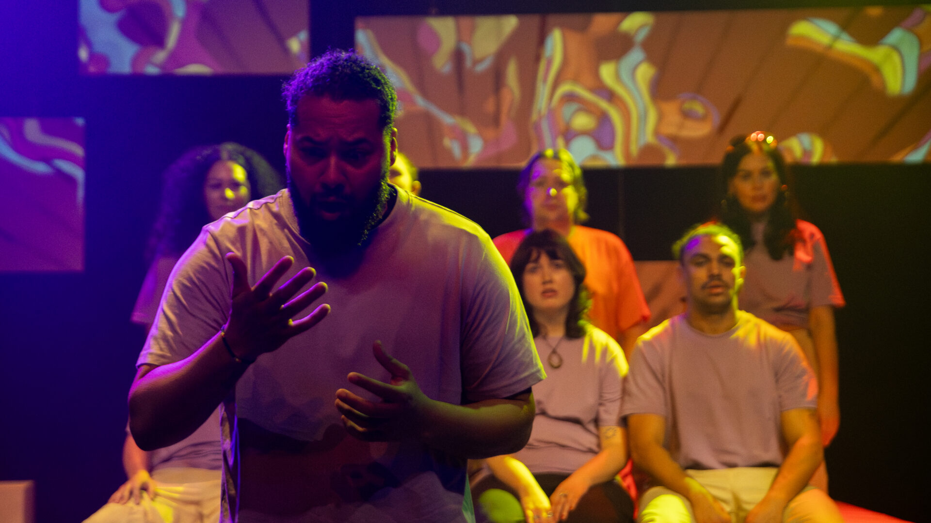 A man stands on stage looking worriedl at his hands, people sit behind him looking at him.